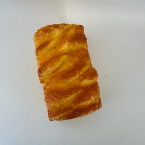 May include: A rectangular piece of food with a golden-brown, textured crust and a light yellow interior. The surface has a wavy pattern, and the edges are slightly uneven. The food item is set against a plain white background.
