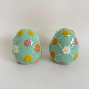 May include: Two egg-shaped salt and pepper shakers. The shakers are a light teal color and decorated with a floral pattern in yellow, orange, pink, and white. The flowers are arranged in a grid pattern with white dotted lines.
