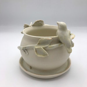 May include: White ceramic planter with a bird perched on a branch, surrounded by leaves. The planter has a matching saucer.