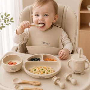 May include: A beige silicone feeding set including a suction bowl, a three-section divided plate, a lidded cup with two handles and a straw, a spoon, and a fork, all personalized with the name Oliver. The set also includes two pacifiers.