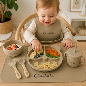 May include: A beige silicone baby feeding set including a bear-shaped divided plate, a bowl, a lidded cup with a straw, a spoon, a fork, and a placemat. Each item is personalized with the name Charlotte in a dark script font.