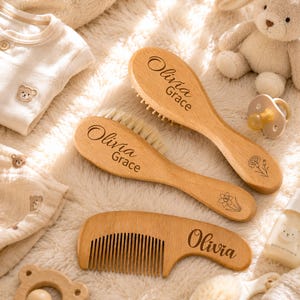 Puede incluir: Dos cepillos de madera con cerdas suaves y un peine de madera, todos con el nombre grabado Olivia Grace u Olivia. Los cepillos incluyen grabados de líneas florales y los artículos están dispuestos sobre una superficie crema suave y texturizada.
