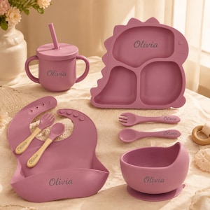 May include: A mauve silicone feeding set personalized with the name Olivia. The set includes a dinosaur-shaped divided plate, a lidded cup with two handles and a straw, a bib with a crumb catcher, a bowl, and two spoons and a fork with wooden handles.