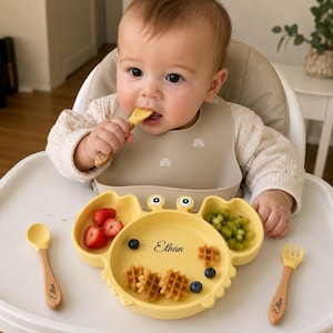Puede incluir: Un plato de silicona amarillo con forma de cangrejo con el nombre Ethan impreso en negro, acompañado de una cuchara y un tenedor de silicona amarilla con mangos de madera grabados con el nombre Ethan, y un babero de silicona beige con arcoíris.
