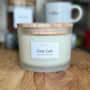Puede incluir: Un tarro de vela de vidrio blanco esmerilado con tapa de madera. La vela está etiquetada como "Cozy Café" y "Hand Poured Coconut Candle" con el texto "Essentially E" sobre la etiqueta.