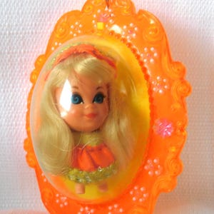 May include: A vintage doll in an orange plastic frame. The doll has blonde hair, blue eyes, and wears an orange dress. The frame is oval-shaped with decorative floral accents and a small loop for hanging.