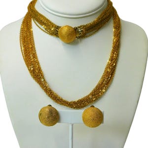 May include: A gold-toned necklace and matching earrings set. The necklace features multiple strands of delicate chains, connected by a large, textured spherical clasp. The earrings are round, textured studs. The jewelry is displayed on a white mannequin.
