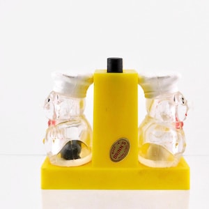 May include: A pair of clear plastic salt and pepper shakers shaped like chefs, with white hats and red bow ties. The shakers are on a yellow base with a red "Made in U.S.A. BRINK'S" label.