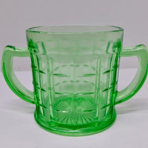 May include: A green glass sugar bowl with two handles. The bowl has a rectangular grid pattern and a circular base with a sunburst design. The glass is a vibrant, translucent green.