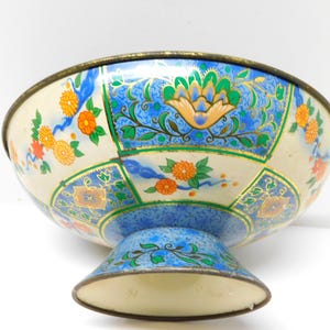 May include: A decorative footed bowl with a cream-colored exterior and intricate floral patterns in blue, orange, and green. The bowl has a gold-colored rim and base, and the design is reminiscent of vintage or antique pieces.