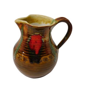 May include: A ceramic pitcher with a handle, featuring a brown base with a red leaf design and green accents. The rim is a light yellow color. The pitcher is likely used for serving beverages.
