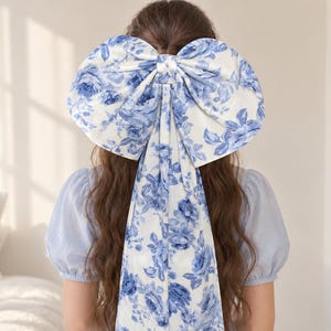 May include: A large hair bow featuring a white fabric base with a blue floral pattern. The bow has two long, wide fabric tails that drape down the back, matching the floral print of the bow. The accessory is secured at the center with a knot.