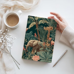 May include: A journal with a jungle-themed cover featuring an elephant, birds, and lush foliage in shades of green, brown, and peach. The journal is held by a person's hand, with a cup of coffee and a pen nearby.
