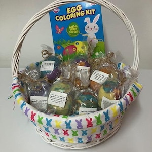 May include: A white wicker basket filled with colorful Easter egg-shaped soaps and an "Egg Coloring Kit" box. The basket's lining features a repeating pattern of colorful bunnies. The soaps are individually wrapped.