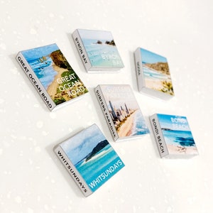 Könnte beinhalten: Eine Sammlung von Miniaturbüchern mit Reise-Themen, die malerische Strand- und Küstenansichten zeigen. Titel sind "Great Ocean Road", "Byron Bay", "Noosa", "Surfers Paradise", "Whitsundays" und "Bondi Beach". Die Bücher haben weiße Ränder.