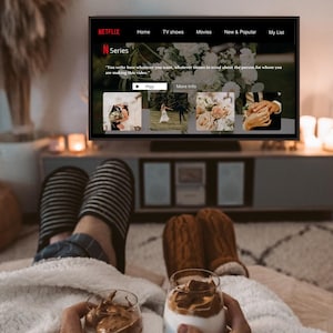 Könnte beinhalten: Eine gemütliche Szene mit einem Netflix-Bildschirm, der ein Hochzeitsvideo zeigt. Zwei Gläser Kaffeegetränke werden von Personen gehalten, die Hausschuhe tragen und sich auf einer weichen Decke entspannen. Kerzen und dekorative Elemente schaffen eine warme Atmosphäre.