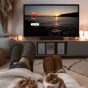 May include: A television screen displays a sunset over water with the text GUESS THE MOVIE, Question, and Answer. Two people hold glasses of whipped coffee while wearing striped and brown cable-knit slippers under a white sherpa blanket.