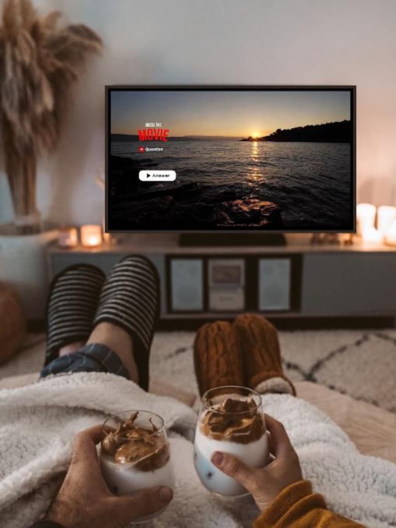 May include: A television screen displays a sunset over water with the text GUESS THE MOVIE, Question, and Answer. Two people hold glasses of whipped coffee while wearing striped and brown cable-knit slippers under a white sherpa blanket.