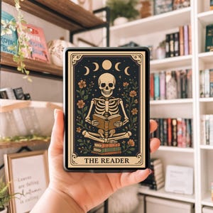 May include: A tarot card featuring a skeleton reading a book, with a dark blue background and floral accents. The card is framed with a gold border and the text "THE READER" is at the bottom. The card is held in a person's hand.