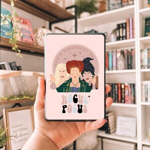 May include: A pink phone case with a graphic of three women and the words "Hocus Pocus" and "House of Sanderson Est. 1693". The case has a black border and is held in a hand. The background includes books and shelves.