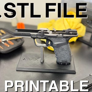 May include: .STL FILE image with a disassembled black handgun on a black stand. The stand is labeled with the text "PRINTABLE CLEANING STAND". A cleaning kit and other gun parts are visible in the background.