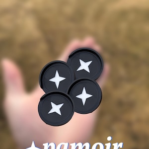 May include: Four black lens caps with a white star design. The lens caps are stacked on top of each other and are being held in a hand. The background is blurred and out of focus. The text "pamoir LABS" is visible in the lower part of the image.