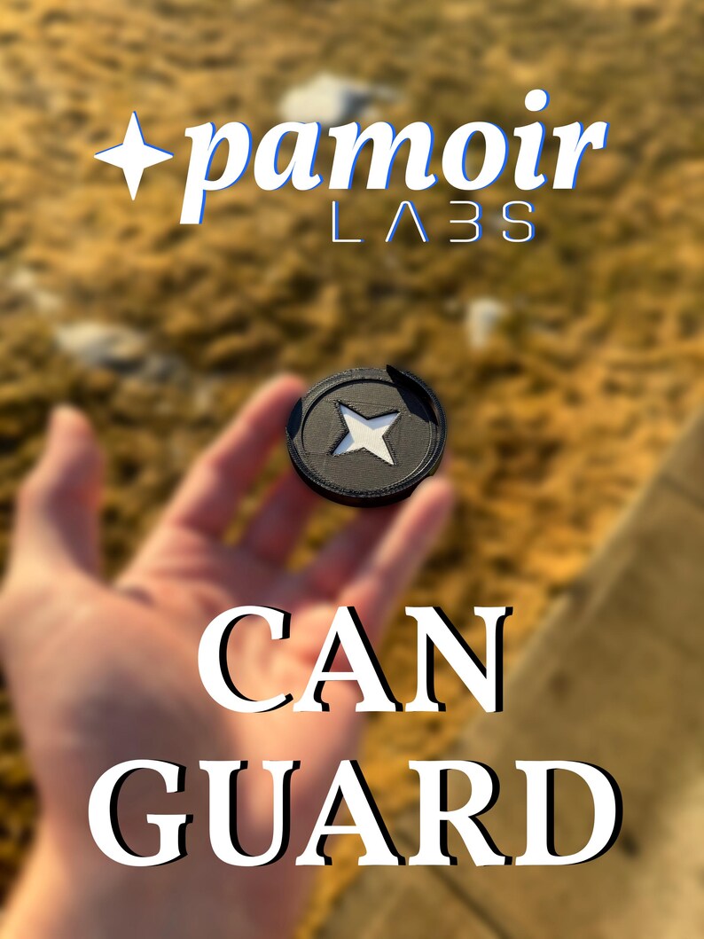 May include: A black can guard with a white star design in the center. The text "pamoir LABS" is at the top, and the words "CAN GUARD" are at the bottom. The background is blurred, with a natural, outdoor setting.