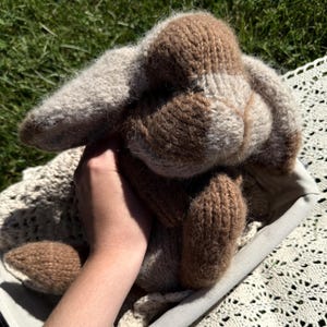 May include: A knitted stuffed rabbit toy featuring a two-tone color scheme of light beige and medium brown yarn. The plush animal has long, floppy ears, a rounded snout, and a soft, textured surface created by the knit stitches throughout its body.
