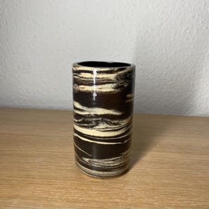 Könnte beinhalten: Eine zylindrische Keramikvase mit einem wirbelartigen Muster in Dunkelbraun und Creme. Das Innere der Vase ist schwarz. Die Vase steht auf einer Holzoberfläche.