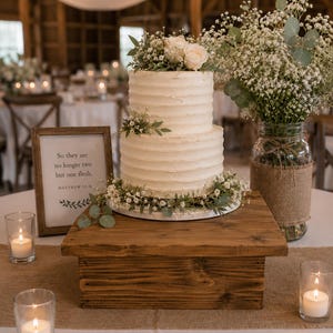 May include: A two-tier white frosted cake topped with white roses and baby's breath, sitting on a square wooden stand. Next to it is a framed sign reading, "So they are no longer two but one flesh. MATTHEW 19:6" and a glass jar filled with flowers.