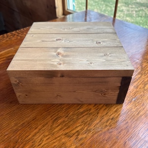 May include: A rectangular wooden box with a light brown finish. The lid is made of several wooden planks. The box is sitting on a wooden table. The box is a natural wood color.