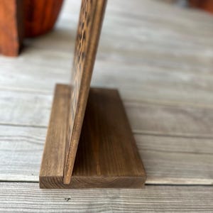 May include: A wooden sign with a dark brown finish, propped up on a matching base. The sign appears to have engraved text. The wood grain is visible, adding texture. The sign is displayed on a wooden surface.