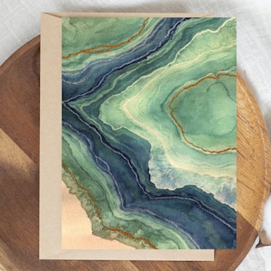 May include: A rectangular greeting card featuring a watercolor-style abstract design of layered agate stone patterns in shades of deep navy blue, sage green, and cream, accented with thin, shimmering copper-colored lines throughout the artwork.