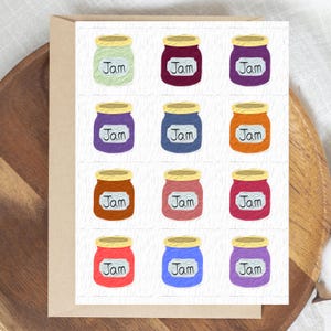 May include: A greeting card featuring a grid of twelve jars of jam. Each jar is a different color, including green, purple, orange, and red, with a gold lid and a label that reads "Jam". The card is on a wooden tray.