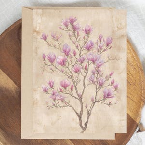 May include: A greeting card featuring a watercolor illustration of a magnolia tree in full bloom. The flowers are a soft pink, set against a vintage-style beige background. The card is displayed on a wooden tray.