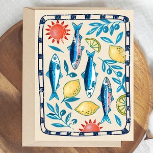 May include: A rectangular greeting card featuring a cream background with a dark blue checkered border. The design includes illustrations of four blue fish, two yellow lemons, two orange suns, and blue olive branches with leaves and fruit.