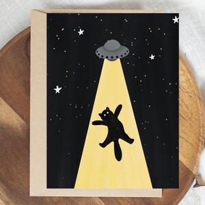 May include: A rectangular greeting card featuring a black background with white stars and a gray UFO. A bright yellow beam of light shines down from the UFO, illuminating a black cat that appears to be floating upward into the spacecraft.