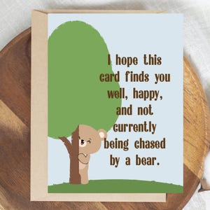 May include: A greeting card with a light blue background, featuring a cartoon bear peeking from behind a tree. The card includes the text: "I hope this card finds you well, happy, and not currently being chased by a bear."