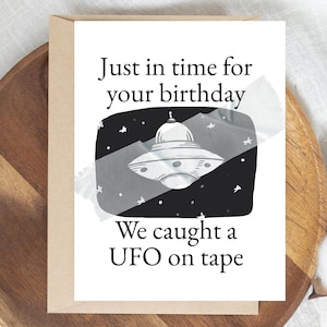 May include: A birthday card with the text "Just in time for your birthday" and "We caught a UFO on tape." The card features a black and white illustration of a UFO, taped to the card. The card is on a wooden surface.