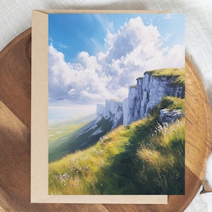 May include: A landscape art print featuring a dramatic cliffside scene under a bright blue sky with fluffy white clouds. The foreground shows a grassy hillside with wildflowers, and the print is set in a brown card.