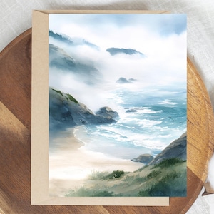 May include: A watercolor art print of a coastal landscape. The artwork depicts a beach with ocean waves, rocky cliffs, and a misty sky. The print is on a beige card, and the overall color palette includes shades of blue, green, and beige.