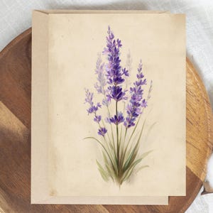 May include: A greeting card featuring a watercolor illustration of lavender. The artwork showcases vibrant purple lavender flowers with green stems and leaves against a beige background. The card is displayed on a wooden tray.