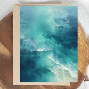 May include: A greeting card featuring a watercolour design of ocean waves in teal, turquoise, and white. The card is inside a brown envelope, resting on a wooden tray.