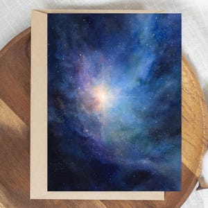 May include: A greeting card with a watercolour painting of a vibrant nebula. The artwork shows a bright, central starburst with radiating lines, surrounded by swirling blues, purples, and hints of orange and green. The card is set in a brown envelope.