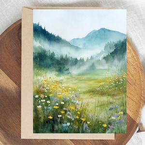 May include: Watercolor landscape art print featuring a serene mountain scene with a meadow of wildflowers. The artwork showcases a valley with lush greenery, yellow, white, and purple flowers, and misty mountains in the background. The print is on a beige card.
