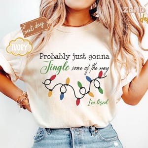 Pode incluir: Camiseta cor marfim com o texto "Probably just gonna Jingle some of the way. I'm tired." e uma sequ&ecirc;ncia de luzes de Natal. A camiseta tem mangas curtas e &eacute; feita de um material macio.