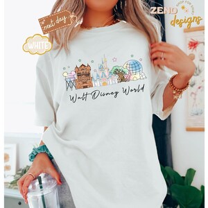 May include: White t-shirt featuring a colorful illustration of the Walt Disney World skyline. The design includes iconic landmarks like Cinderella Castle and Spaceship Earth, with the words "Walt Disney World" in a script font.