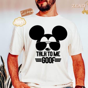 Puede incluir: Una camiseta de manga corta color marfil con un gr&aacute;fico negro de la silueta de un rat&oacute;n con gafas de sol. Debajo del gr&aacute;fico, el texto dice TALK TO ME GOOF con l&iacute;neas horizontales a ambos lados, centrado en la parte frontal.