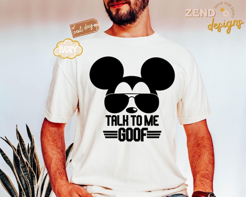 Puede incluir: Una camiseta de manga corta color marfil con un gr&aacute;fico negro de la silueta de un rat&oacute;n con gafas de sol. Debajo del gr&aacute;fico, el texto dice TALK TO ME GOOF con l&iacute;neas horizontales a ambos lados, centrado en la parte frontal.
