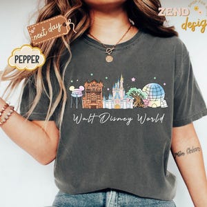 May include: Dark gray t-shirt featuring a colorful illustration of Walt Disney World landmarks. The design includes the Tower of Terror, Cinderella Castle, and Spaceship Earth, with the words "Walt Disney World" below.